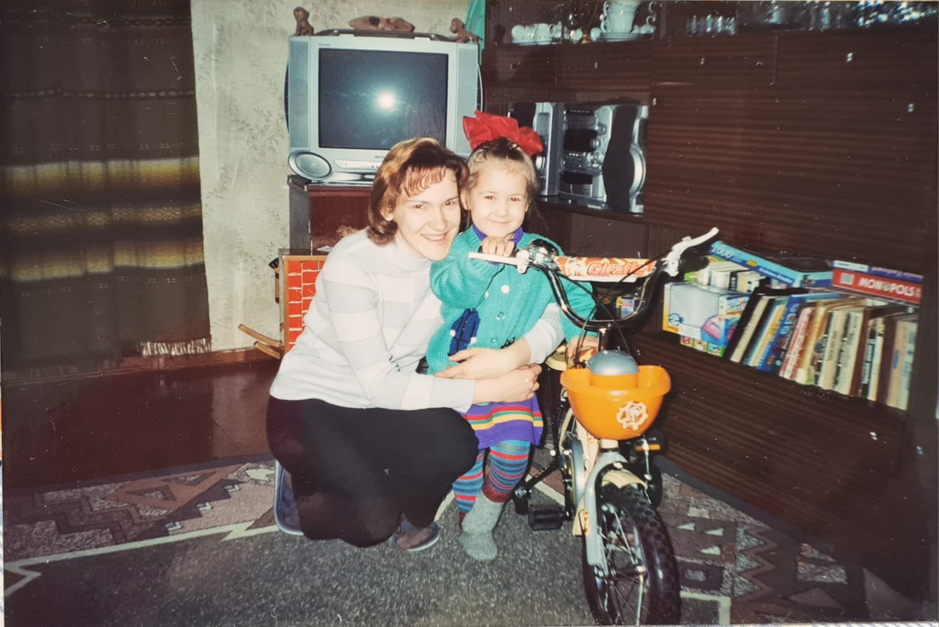 Mamma un meita abas apskāvušās, blakus jauns velosipēds, fonā brūna padomju sekcija, vecs televizors