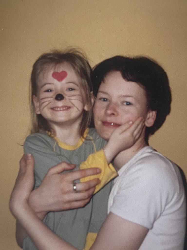 Mamma tur rokās meitu, kurai us sejas uzzīmētās kaķa ūsas un sirsniņa uz pieres